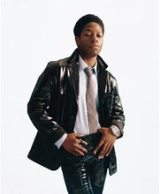 A young man wearing a shiny black leather jacket, white dress shirt, and patterned tie, posed against a plain white studio background.