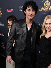 A man in a black leather blazer and black shirt stands next to a blonde woman in a black sequined dress in front of a dark event backdrop with logos.