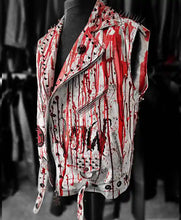 A sleeveless moto-style vest displayed on a black mannequin torso, featuring a white base covered in bold red paint-splatter and black distressed graphics, with metal studs, zippers, and a pointed collar.