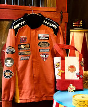 An orange and black racing jacket with multiple sponsor patches is displayed on a hanger next to a matching orange, white, and red tote bag decorated with racing patches, set against a rustic wooden background.