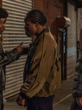 A man in a brown suede bomber jacket with black leather trim stands in a street setting at night, looking to the side while another person is partially visible on the left.