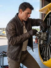 A man in a brown leather jacket and gloves works on a large aircraft engine inside an airfield hangar.