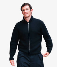 A man wearing a black ribbed zip-up sweater with a stand collar and matching dark trousers, photographed against a plain light gray background.