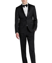 A man wearing a black tuxedo with satin peak lapels, a white dress shirt, and a black bow tie, photographed against a plain light gray studio background.