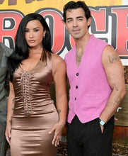 Demi Lovato and Joe Jonas posing together at an event, with Demi wearing a metallic bronze satin halter dress and Joe wearing a pink sleeveless vest over a white undershirt.