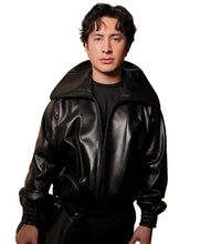 A man wearing a black leather jacket with a high collar over a black t-shirt, standing against a textured stone wall background.
