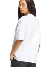 A woman wearing a white short-sleeve top and black pants, shown from a side-back angle against a light gray studio background.