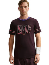 A man wearing a dark maroon Nike t-shirt with 'VAMOS' printed across the chest in white and pink, featuring a pink and blue striped collar and sleeve cuffs, paired with matching maroon shorts against a light gray background.