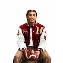 A young man sitting and holding a football, wearing a maroon and white varsity jacket with a large gold letter 'H' on the chest, brown pants, and a white t-shirt underneath, set against a solid white background.