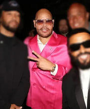 A man wearing a bright pink satin double-breasted blazer, white dress shirt, and sunglasses, making a peace sign. He is surrounded by other men in dark clothing in a dimly lit setting.