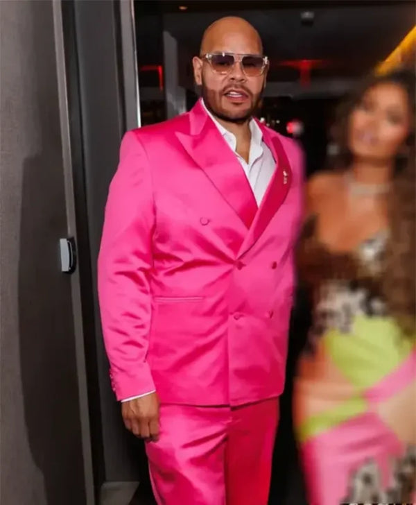 A man wearing a bright hot pink double-breasted suit with a white shirt and sunglasses, standing in a dimly lit indoor setting.