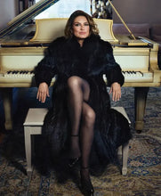 A woman seated on a bench in front of a cream-colored grand piano, wearing a voluminous black fur coat, sheer black tights, and black heels in an indoor room with a patterned rug.