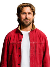 A man with light brown hair and a beard wearing a red button-up jacket over a white t-shirt, standing against a dark background.