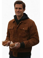 A man wearing a brown suede jacket over a black hoodie, holding two hot dogs against a plain white background.