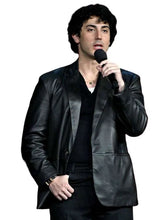 A man with dark curly hair wearing a black leather jacket over a black shirt, holding a black microphone against a plain white background.
