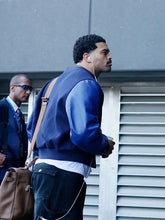 A man wearing a blue leather-look bomber jacket, dark cargo pants, and a brown shoulder bag walks outdoors next to a gray building with louvered windows. Another man in sunglasses is partially visible in the background.