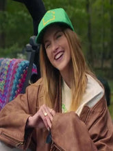 A woman smiling while wearing a green baseball cap with a yellow graphic, a brown jacket, and a cream shirt, seated outdoors.