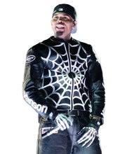 A man wearing a black leather jacket with a white spiderweb pattern on the front, black gloves, and a black baseball cap, standing in front of a blurred crowd.