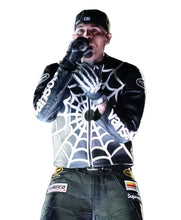 A performer on stage wearing a black and white spiderweb-patterned jacket with 'Vans' branding, black gloves, and a black cap, holding a microphone in front of a blurred crowd.