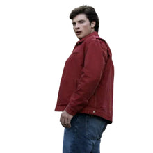 A man wearing a red jacket and blue jeans standing outdoors in a wooded setting.