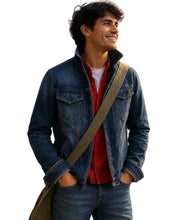 A man wearing a blue denim jacket over a red button-up shirt, standing outdoors in a rustic village setting with colorful papel picado banners on the wall behind him.
