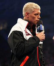 A man with platinum blonde hair wearing a black, white, and red zip-up jacket holds a microphone with a blue and white logo, set against a dark blue background.