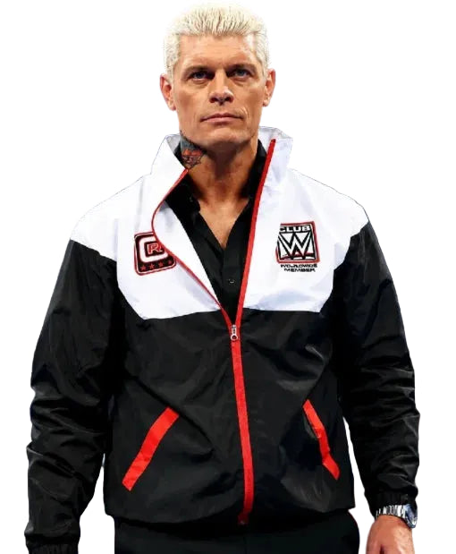 A man with platinum blonde hair wearing a black and white zip-up jacket with red trim and WWE patches, standing against a dark blue background.