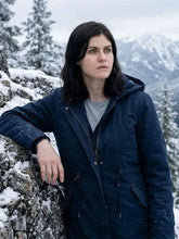 A woman wearing a dark navy hooded winter parka over a gray shirt, standing outdoors in a snowy mountain landscape.