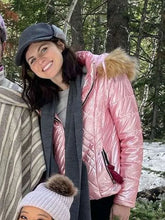 A woman outdoors in a snowy forest wearing a shiny pink quilted winter jacket with a faux-fur trimmed hood, a dark gray scarf, a dark gray flat cap, and a light pink knit beanie with a brown pom-pom.