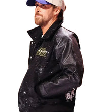 A man wearing a black glittery varsity-style jacket with a gold 'Star Wars' logo on the chest, a blue and white baseball cap, and a white undershirt, set against a warm, blurred background.