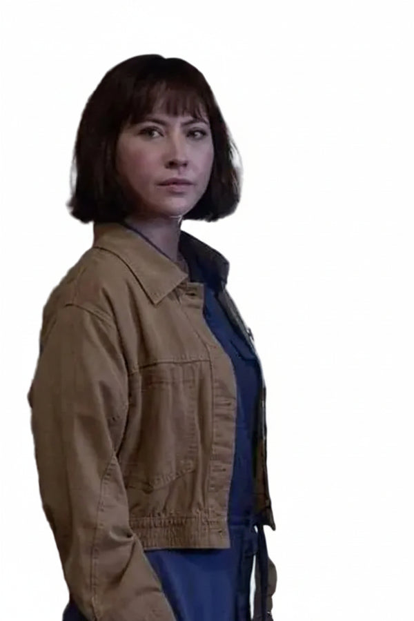 A woman with a short dark bob haircut wearing a tan utility jacket over a blue top, standing in an indoor setting with ornate wooden details. Another person is partially visible on the right.