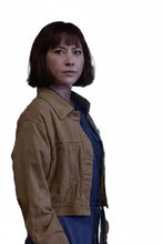 A woman with a short dark bob haircut wearing a tan utility jacket over a blue top, standing in an indoor setting with ornate wooden details. Another person is partially visible on the right.