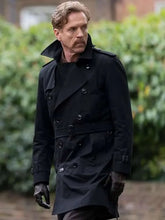 A man wearing a black double-breasted trench coat, black shirt, black gloves, and brown leather boots, standing outdoors in front of a blurred green hedge.
