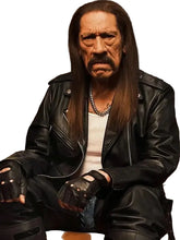 A man with long dark hair and a mustache wearing a black leather motorcycle jacket, black leather pants, fingerless gloves, and a silver chain necklace, seated in a dimly lit room.