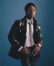 A young man wearing a shiny black leather jacket, white dress shirt, and patterned tie, standing against a solid blue studio background.