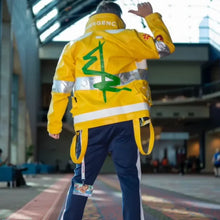 A person shown from behind wearing a bright yellow emergency worker jacket with reflective silver stripes and a large green symbol on the back, paired with navy pants, in an indoor public space.