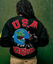 A person shown from behind wearing a black varsity-style jacket with a large back graphic featuring red 'USA' lettering, a black cat, a blue-and-green globe, and red 'DENY ALL' text, against a green textured background.