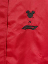Close-up of a red jacket or fabric panel with a black embroidered Mickey Mouse head, an X mark, and an F1 logo, indicating a collaboration.