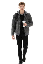 A young man wearing a dark gray washed denim jacket over a black hoodie and black pants, carrying a black backpack and holding a black coffee cup, isolated on a white background.