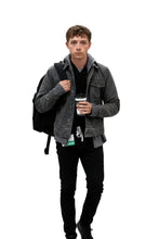 A young man wearing a dark gray washed denim jacket over a black hoodie and black pants, carrying a black backpack and holding a black coffee cup, isolated on a white background.