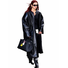A woman modeling a long, oversized black faux-leather coat with a glossy finish, styled with black pants, black boots, black sunglasses, and carrying a black handbag against a plain white background.