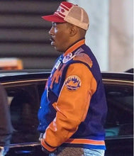 A man in profile wearing a blue and orange varsity jacket with a red and beige trucker hat, standing near a car at night.