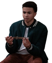 A young man wearing a dark green varsity jacket over a white t-shirt and brown pants, holding a small wooden object in his hands while looking off-camera in a dimly lit indoor setting.