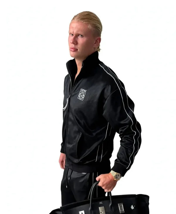 A blonde man wearing a black satin track jacket with white piping and a white crest logo on the chest, paired with black track pants, holding a black handbag against a white background.