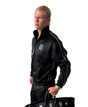 A blonde man wearing a black satin track jacket with white piping and a white crest logo on the chest, paired with black track pants, holding a black handbag against a white background.