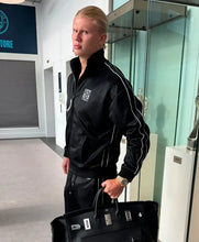 A man wearing a black satin track jacket with white piping and a crest on the chest, standing indoors and holding a black luxury handbag.