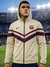 A man wearing a cream-colored FC Barcelona track jacket with dark purple stripes and a navy blue Nike swoosh, standing on a soccer field with stadium lights in the background.