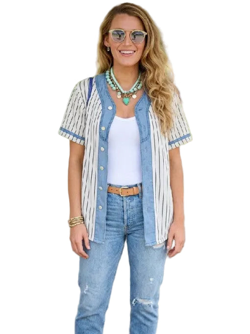 A woman wearing a light blue denim button-up shirt with vertical white stripes, styled over a white tank top and light-wash jeans, standing outdoors under a covered walkway.