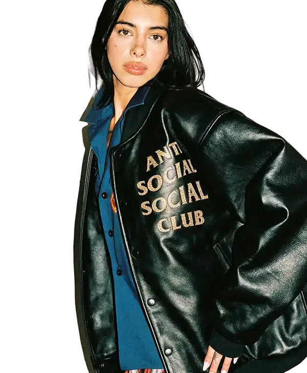 A model wearing a black leather varsity-style jacket with 'ANTI SOCIAL SOCIAL CLUB' embroidered in gold on the chest, layered over a blue button-up shirt against a plain white background.