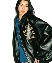 A model wearing a black leather varsity-style jacket with 'ANTI SOCIAL SOCIAL CLUB' embroidered in gold on the chest, layered over a blue button-up shirt against a plain white background.